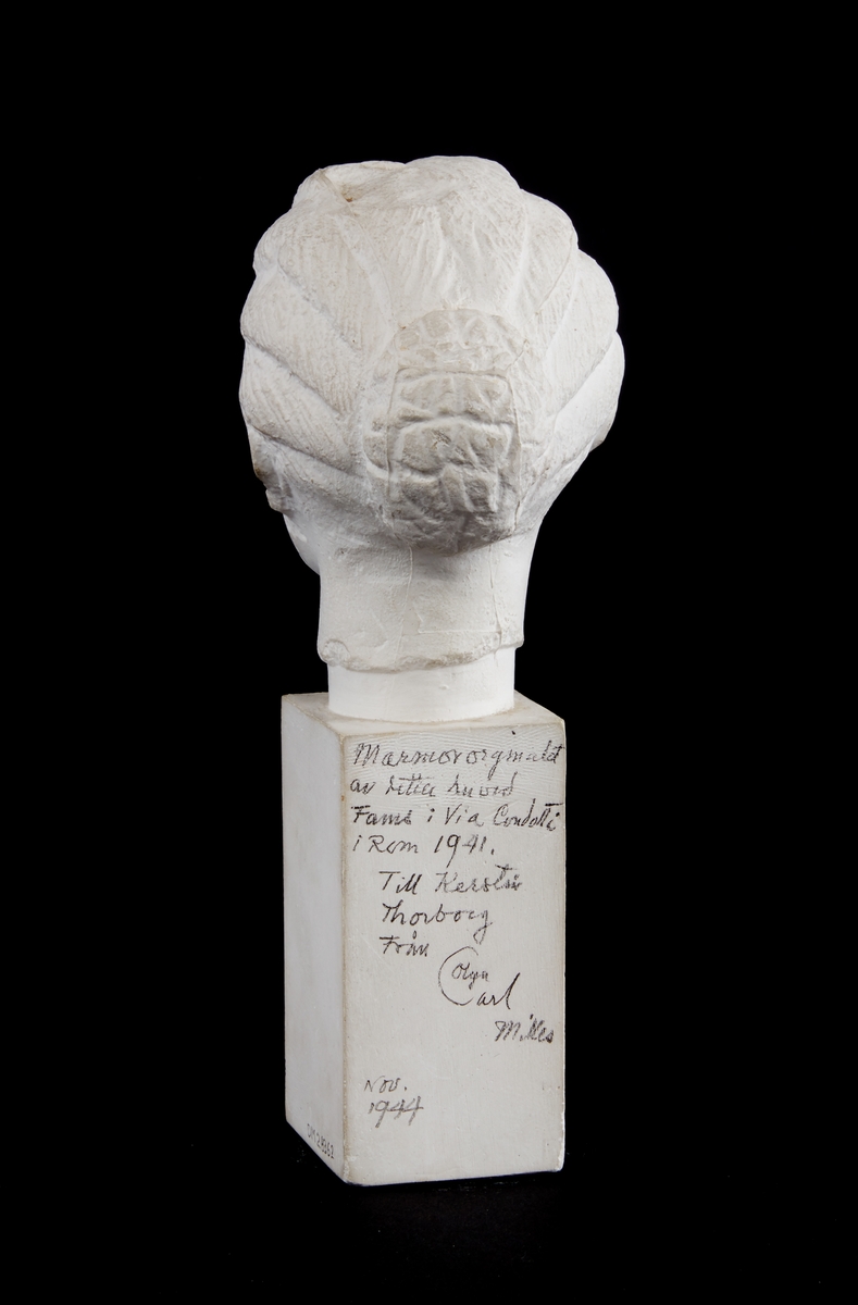 Skulptur, gipsbyst. Hög sockel med kvadratiskt tvärsnitt. Flickhuvud med håret i en knut i nacken. Text med blyerts på baksidan: "Marmororiginalet av detta huvud fanns i Via Condotti i Rom 1941. Till Kerstin Thorborg från Olga, Carl Milles nov. 1944".