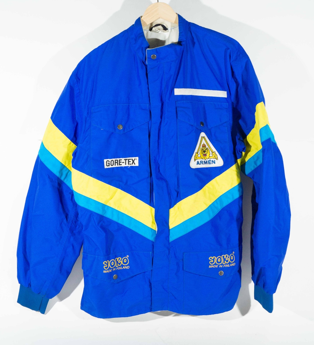 Mörkblått MC-ställ med jacka och byxor med mellanblå och gula detaljer.  På vänster sida av bröstet på jackan "Gore-Tex"  och höger sida ett trekantigt märke "Armén". På ficklocken "YOKO Made in Finland". På ryggen och på byxorna "Arméns Lejon" och "Gore-Tex".