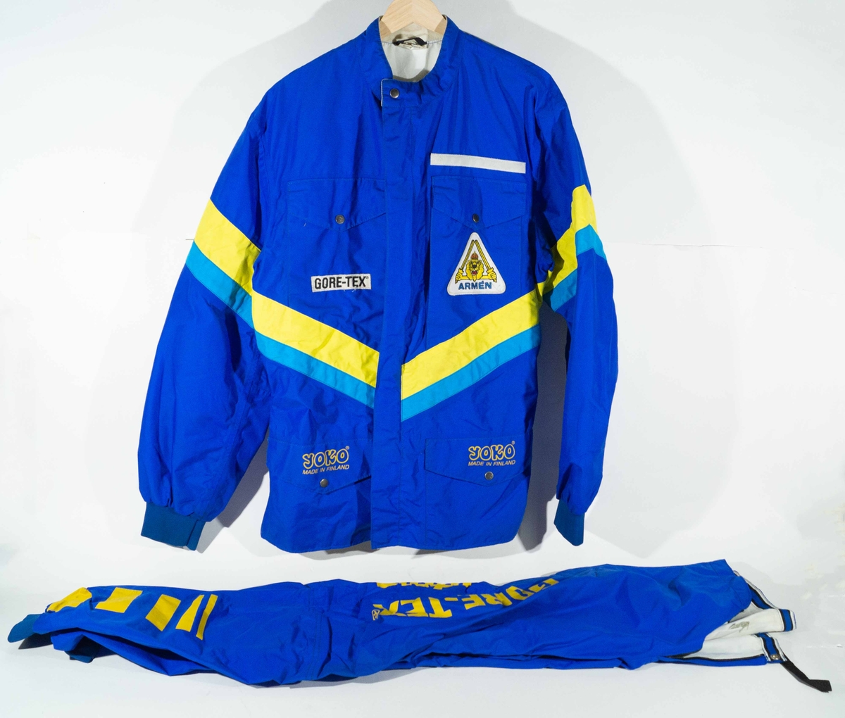 Mörkblått MC-ställ med jacka och byxor med mellanblå och gula detaljer.  På vänster sida av bröstet på jackan "Gore-Tex"  och höger sida ett trekantigt märke "Armén". På ficklocken "YOKO Made in Finland". På ryggen och på byxorna "Arméns Lejon" och "Gore-Tex".