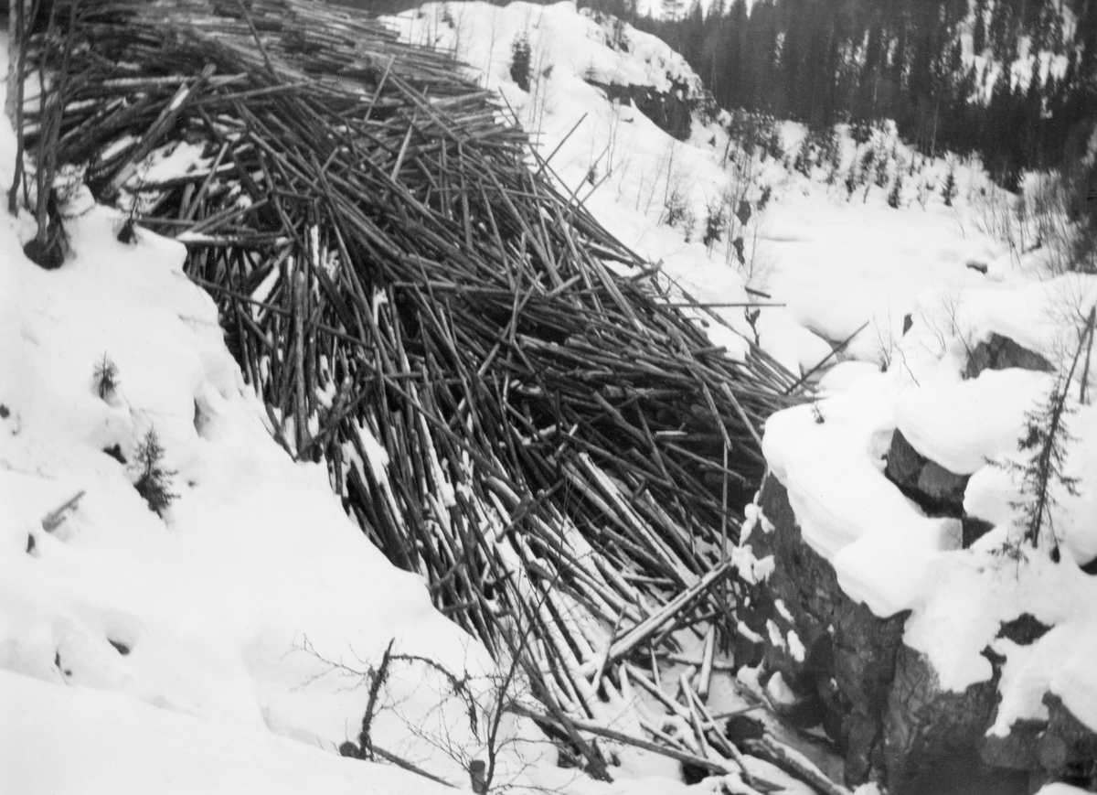 Tømmer som er veltet utfor en bratt snødekt skråning mot et is- og snødekt vassdrag. Fotografiet er fra Torestronget i elva Hovda, ei sideelv på vestsida av Glomma som danner kommunegrense mellom Åmot og Stor-Elvdal i Østerdalen (Hedmark). Bildet er sannsynligvis tatt for å dokumentere en leveranse som var problematisk for fløterne som, når vårflommen kom, skulle få elva til å bære stokkene videre mot hovedvassdraget, Glomma. Her måtte fløterne klatre oppover en bratt elveskrent for å få tømmeret ned i det rennende vannet. Dette var besværlig, tidkrevende og ikke helt ufarlig. I et trangt elvegjel som Torestronget var det dessuten betydelig fare for at tømmeret kunne sette seg fast. Det betydde mer farlig arbeid. Bildet ble tatt i 1938. Dette året var det merket og innmeldt 12 595 tømmerstokker til fløting i Hovda. I tillegg skulle fløterne forsøke å få ut hele 21 690 stokker som lå igjen fra året før. Det greide de nesten. Til tross for leveranser som den vi ser på dette fotografiet var det bare 48 stokker som lå igjen da fløtinga i dette vassdraget ble avsluttet i 1938.