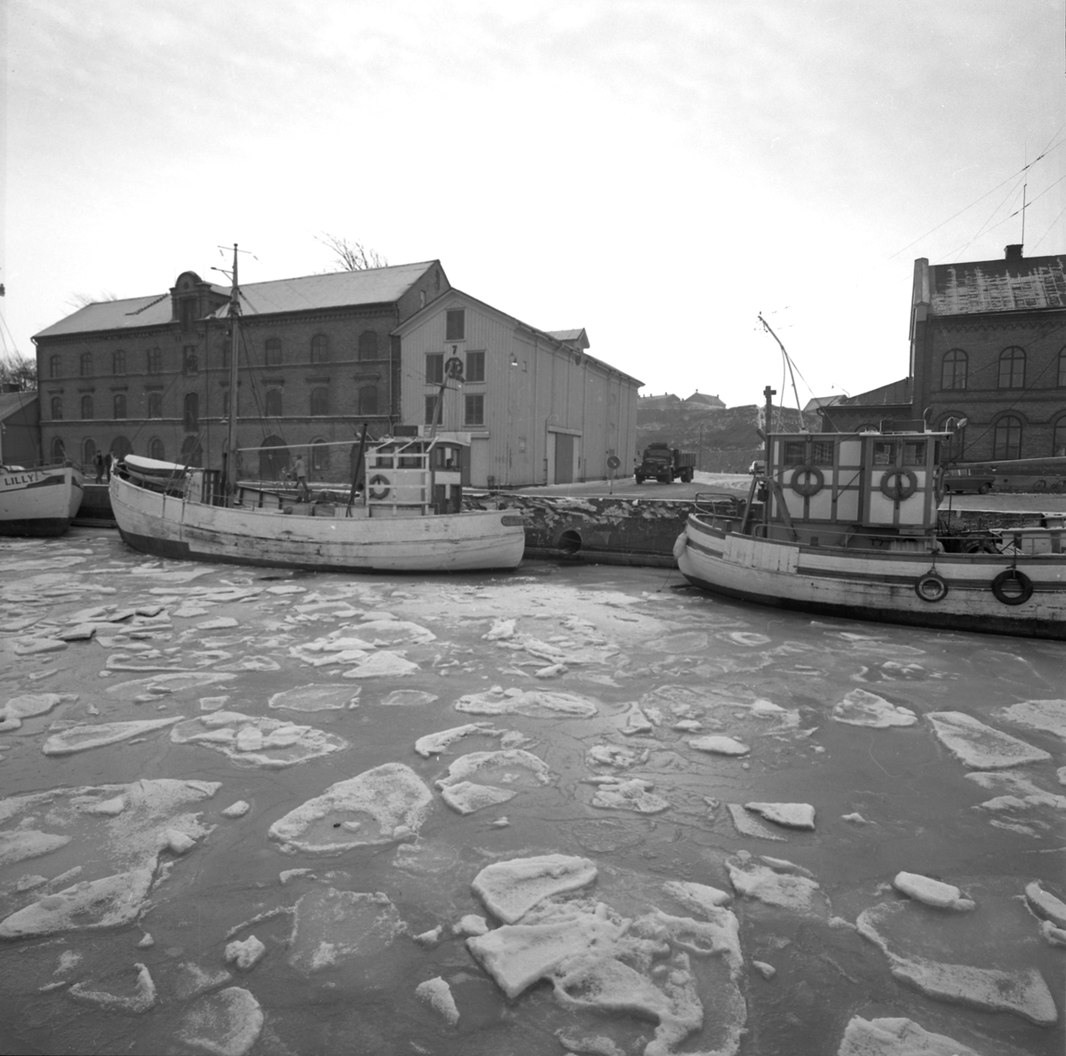 Tidningsartikel med rubriken "Fyra nyårsbåtar i Varbergs hamn" publicerad i Hallands Nyheter 1970-01-02. Till vänster ses hamnmagasinet från 1874 med Lundqvists trämagasin och till höger tullhuset. Fiskebåtar ligger vid kajen och en lastbil står mellan husen.
(Bilderna inlagda i omvänd ordning mot id-numren)