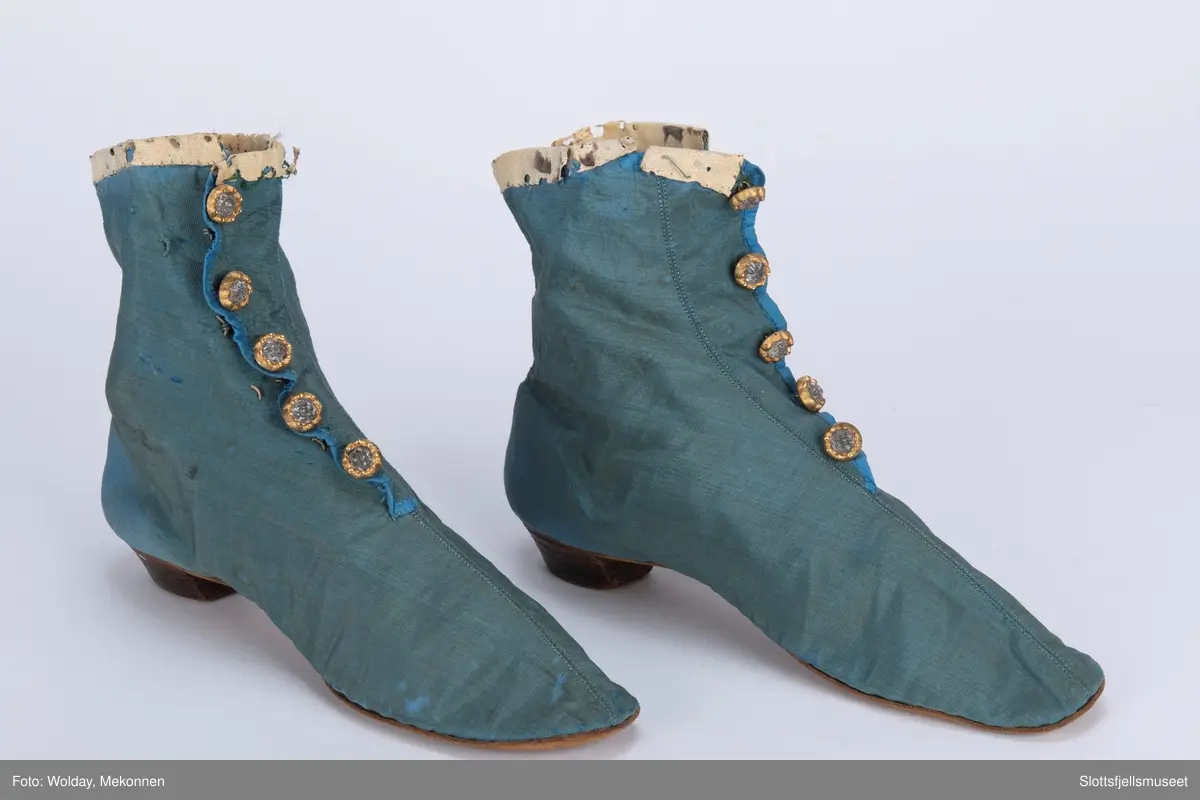 Et par håndsydde støvler. Blå silkerips, stikning langs midten. Innvendig hvitt lerretsfôr og snøring. Lukkes med hekter. Fem pynteknapper. Såle av skinn.