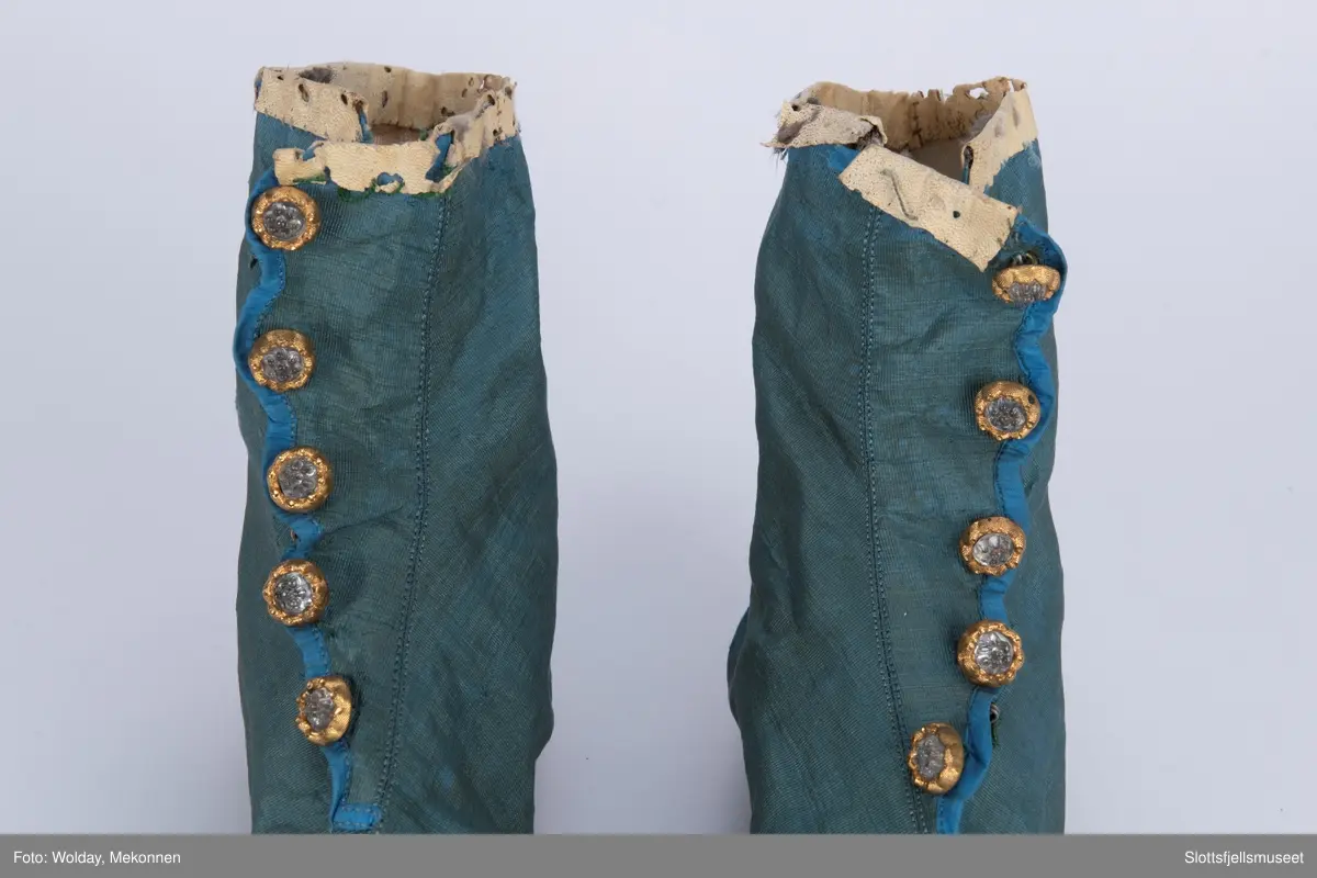 Et par håndsydde støvler. Blå silkerips, stikning langs midten. Innvendig hvitt lerretsfôr og snøring. Lukkes med hekter. Fem pynteknapper. Såle av skinn.