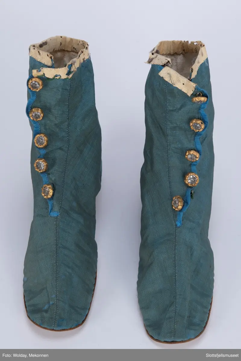 Et par håndsydde støvler. Blå silkerips, stikning langs midten. Innvendig hvitt lerretsfôr og snøring. Lukkes med hekter. Fem pynteknapper. Såle av skinn.