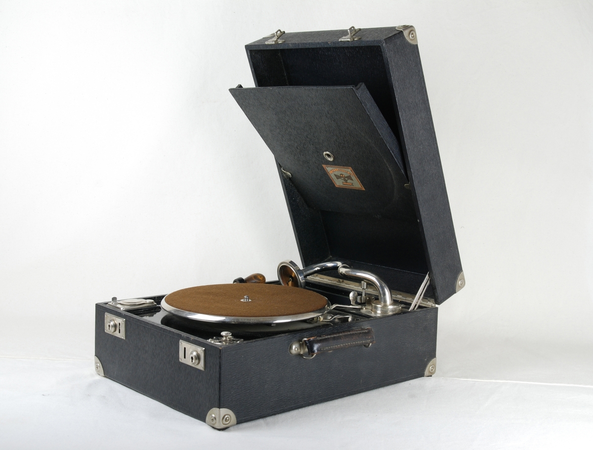 Resegrammofon i svart trälåda. Stor, rund tallrik med knopp i mitten, klädd med brun filt. Övre hörnet: metallarm, rörlig från mitten med rund mellanstor tallrik, där stiftet är fäst. Metallreglage för stängning av locket. Nedre högra hörnet: metallplatta, utsirad med spetsar både uppåt och nedåt. På mitten skruv för att ändra varvtal, under en pil samt siffror 90, 78, 50. Nedre vänstra hörnet: rektangulär metallplatta med öppningsbart lock samt liten skål med stift i. Utefter vänster sida fack för vev. Veven är vinklad med droppformat handtag, i andra ändan gängad för fastsättning. I locket en öppningsbar lucka med plats för skivor. Utsidan har metallhörn med tre nitar i varje, på kortsidan två snäpplås.