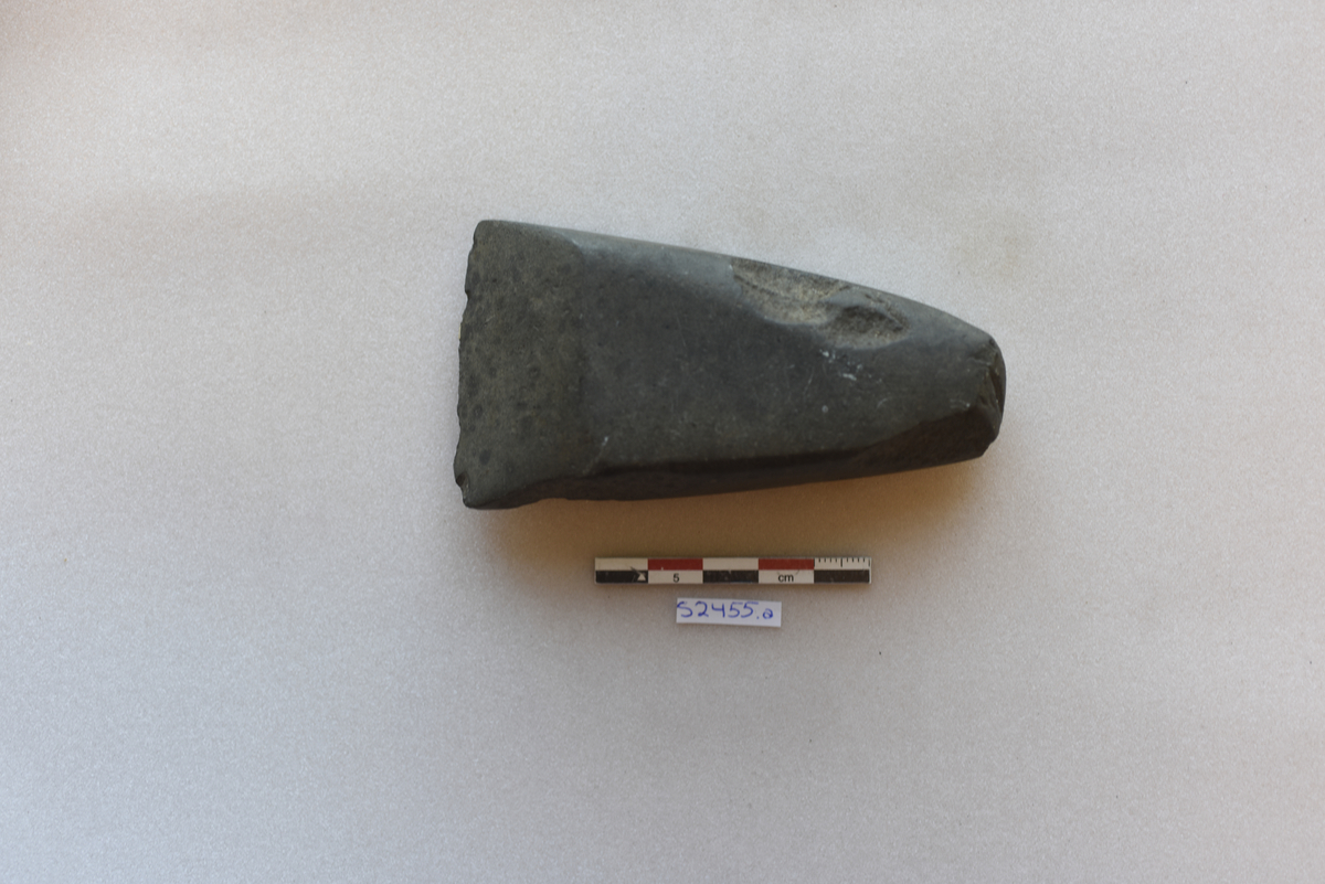 "2455 a. Kile af sleben grønsten, lignende R. 14. Den er 9,8 cm lang, 5,2 cm bred langs eggen og 2,2 cm bred over banen."  Vestlandsøks av grønnstein, avsmalnende mot nakken. Øksa er skadd i eggen, på begge smalsider og i nakken.