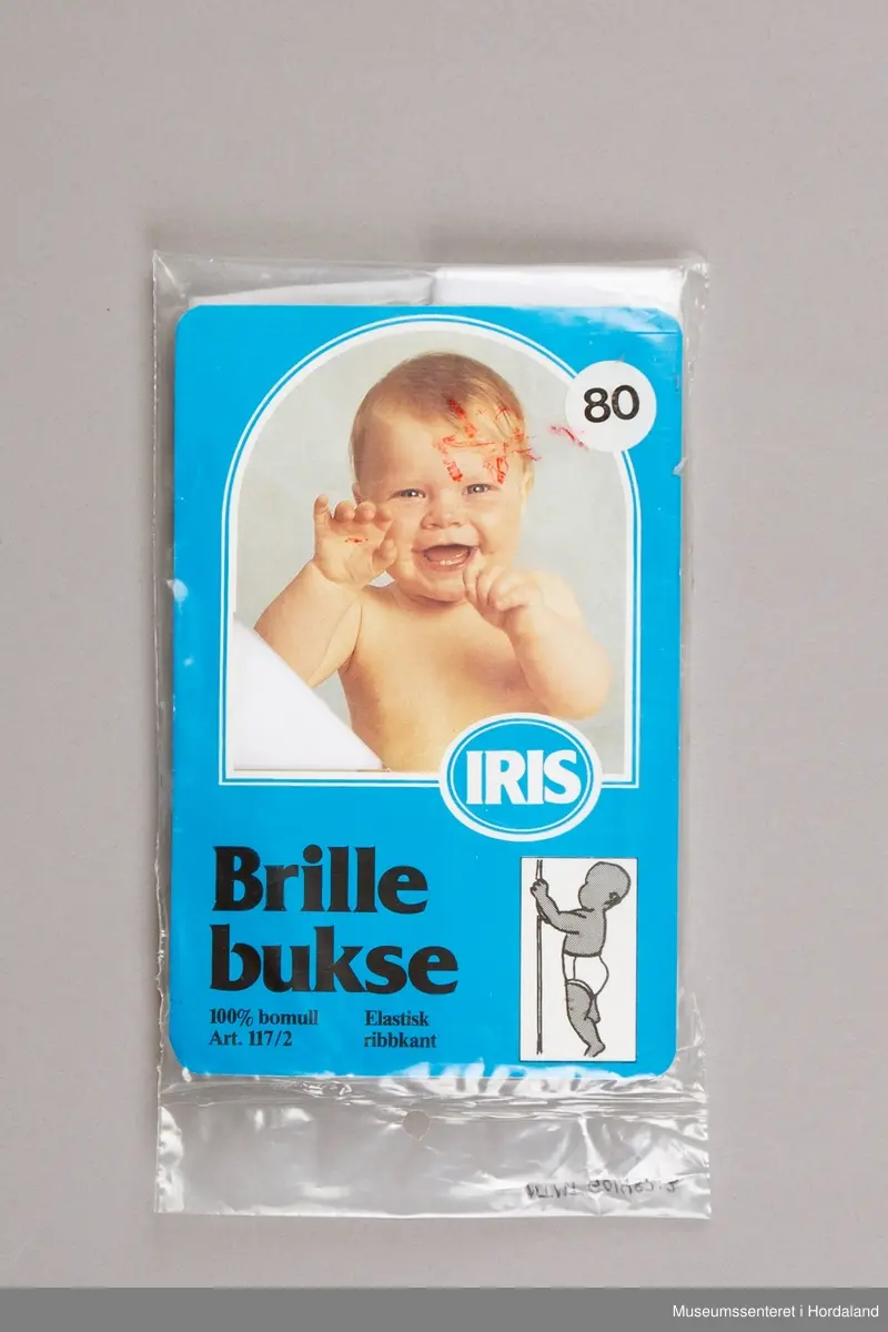 Kvit Iris brillebukse pakka i plastemballasje, med produktinformasjon. Artikkel nummer 117/2, med elastisk ribbkant. Produktinformasjonen som ligg ved gir kvalitetsgaranti, og informasjon om korleis ein kan levere tilbake plagg med feil.