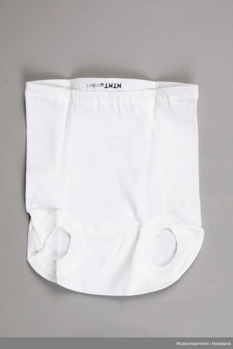 Kvit Iris brillebukse pakka i plastemballasje, med produktinformasjon. Artikkel nummer 117/2, med elastisk ribbkant. Produktinformasjonen som ligg ved gir kvalitetsgaranti, og informasjon om korleis ein kan levere tilbake plagg med feil.