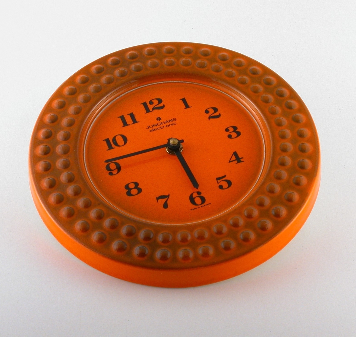 Cirkelrund orange klocka med svarta siffror och visare. Runt om framsidans ytterkant 2 st rader med utbuktande prickar, brun transparent glasyr ovanpå den orangea. Baksidan vitglaserad.