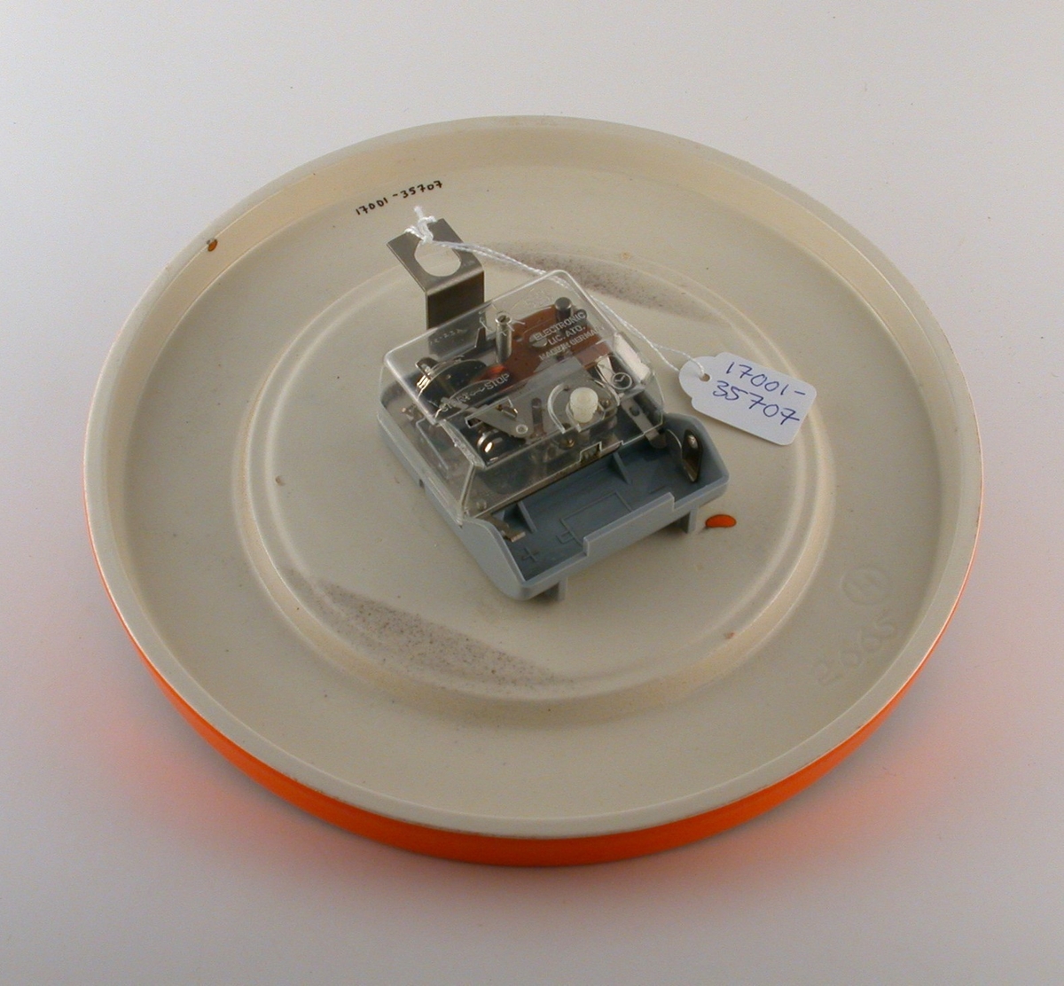 Cirkelrund orange klocka med svarta siffror och visare. Runt om framsidans ytterkant 2 st rader med utbuktande prickar, brun transparent glasyr ovanpå den orangea. Baksidan vitglaserad.