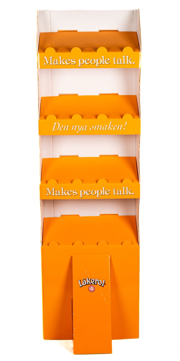 Reklamställ i papp, orange och vitt. Fyra fack för reklamartiklar. Tryckt text: Läkerol Make people talk.