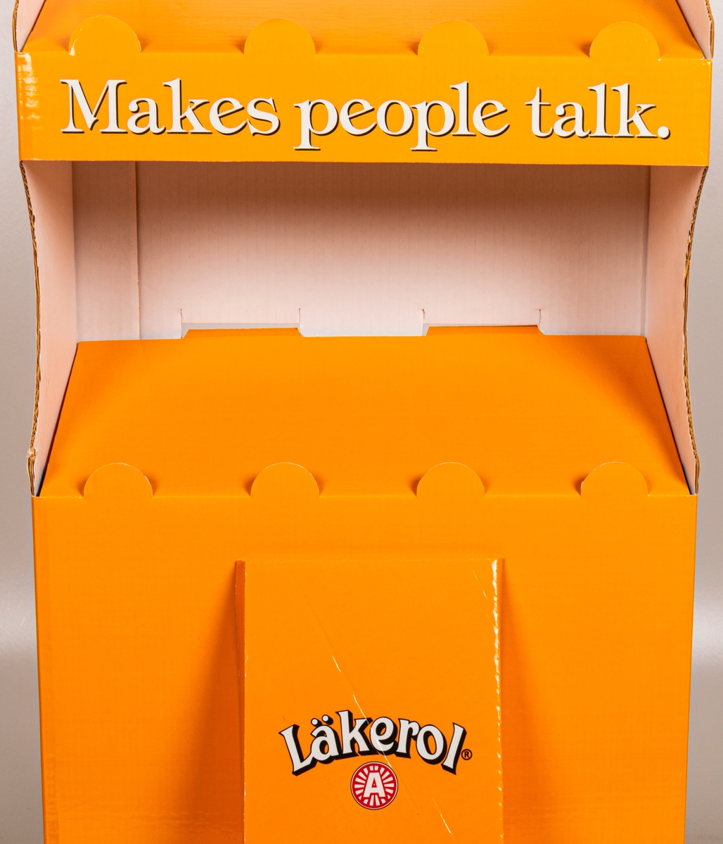 Reklamställ i papp, orange och vitt. Fyra fack för reklamartiklar. Tryckt text: Läkerol Make people talk.