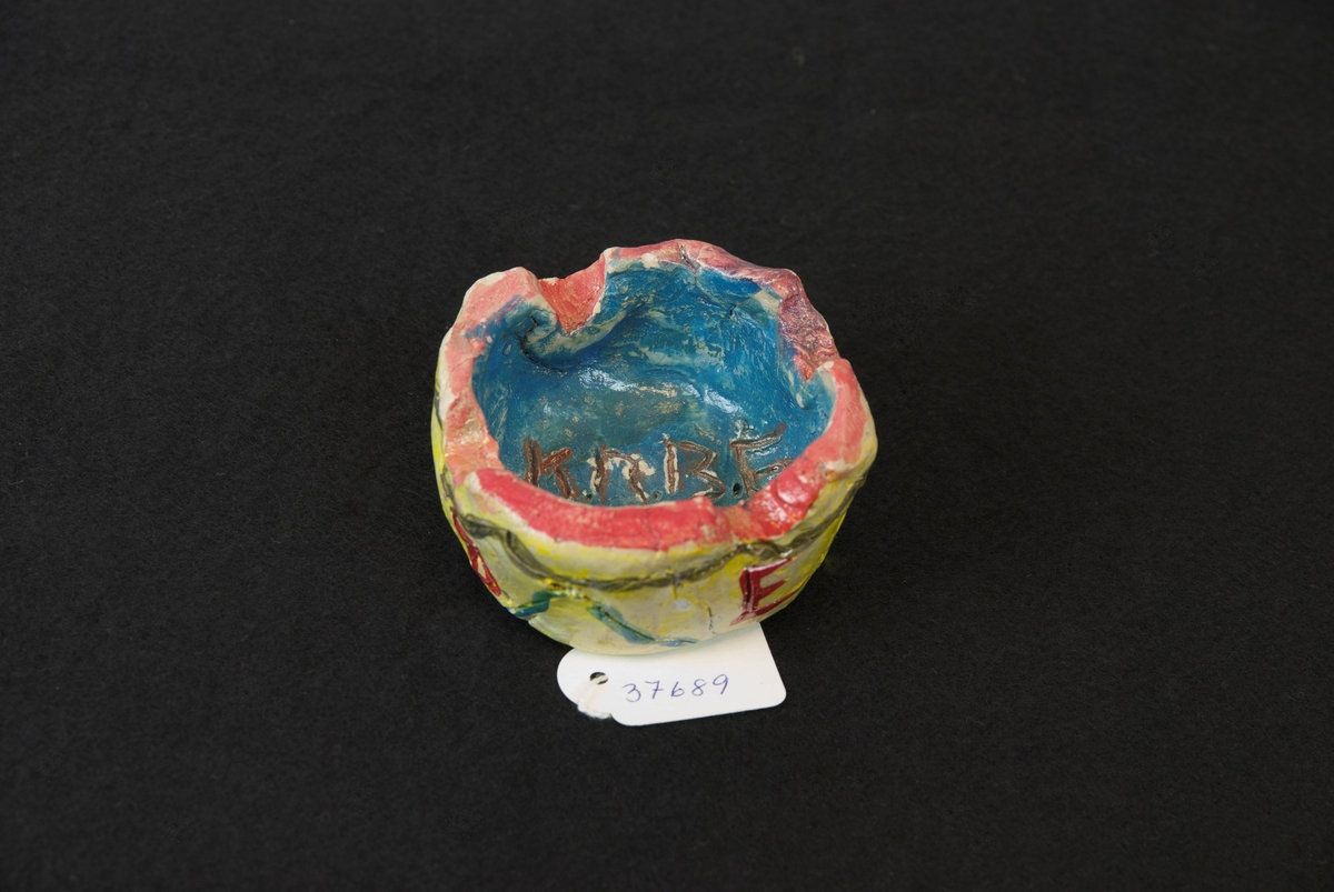 Askfat i lera. Rund form med ca 3,5 cm höga kanter, 4 nedsänkningar i kanten för att lägga cigaretten i.
Dekorationsmålad i gul bottenfärg utvändigt med zickzack-ränder i grönt samt bokstäver K R B E i rött.
 Röd överkant, blå invändigt med bokstäver i rött, K R B E 1941 inristat och målat i brun färg i botten.
Troligtvis skolarbete.