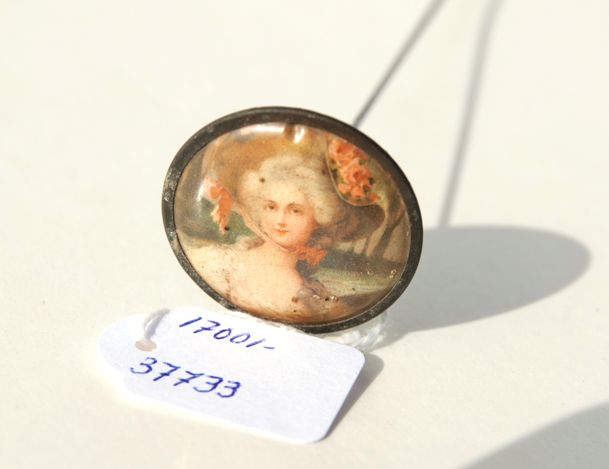 Hattnål i metall. Rund knapp med bild på dam med vitt hår, stor hatt med blommor, fäst mot knappen med smal kant av metall. Guldfärgad baksida med metallsläpp. Lång smal nål för fastsättning i hatten.