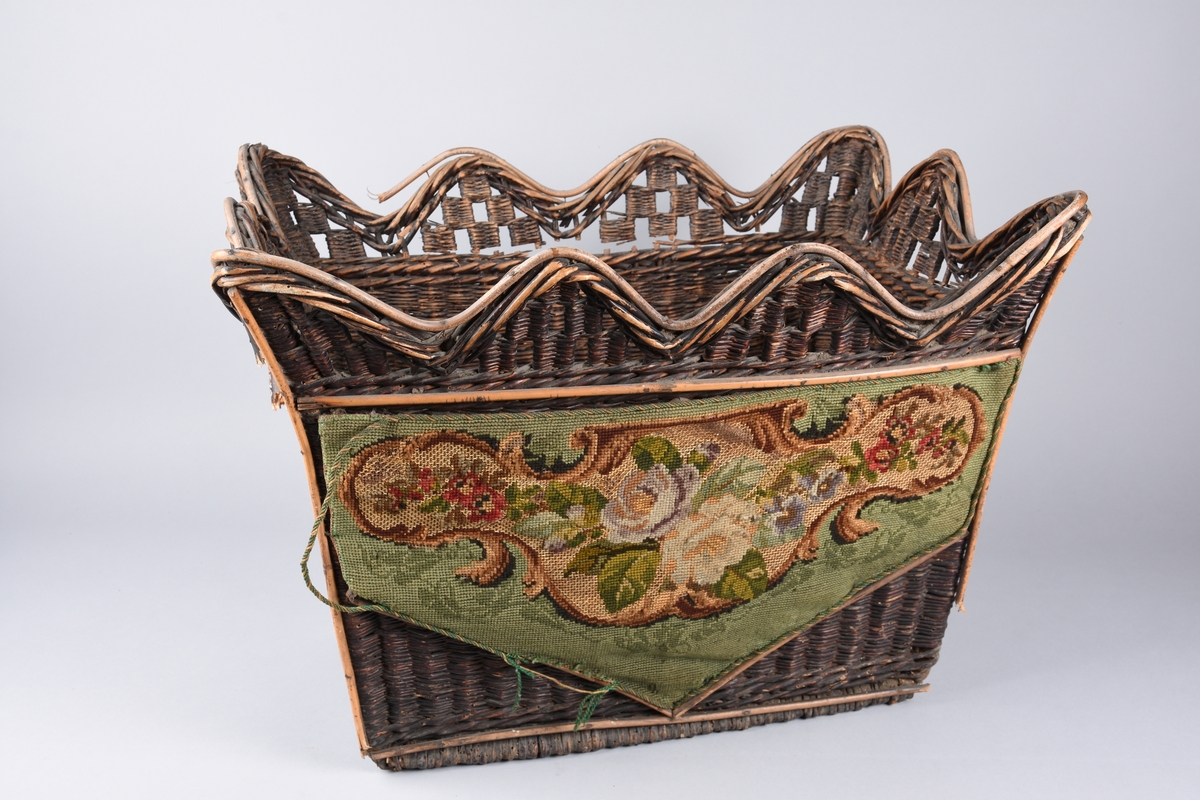 Flettet kurv av vidje med avstivere på tverrsidene. 
Brunmalt. Den ene langsiden er pyntet med broderi i ullgarn. Blomstermønster mot grønn bakgrunn.