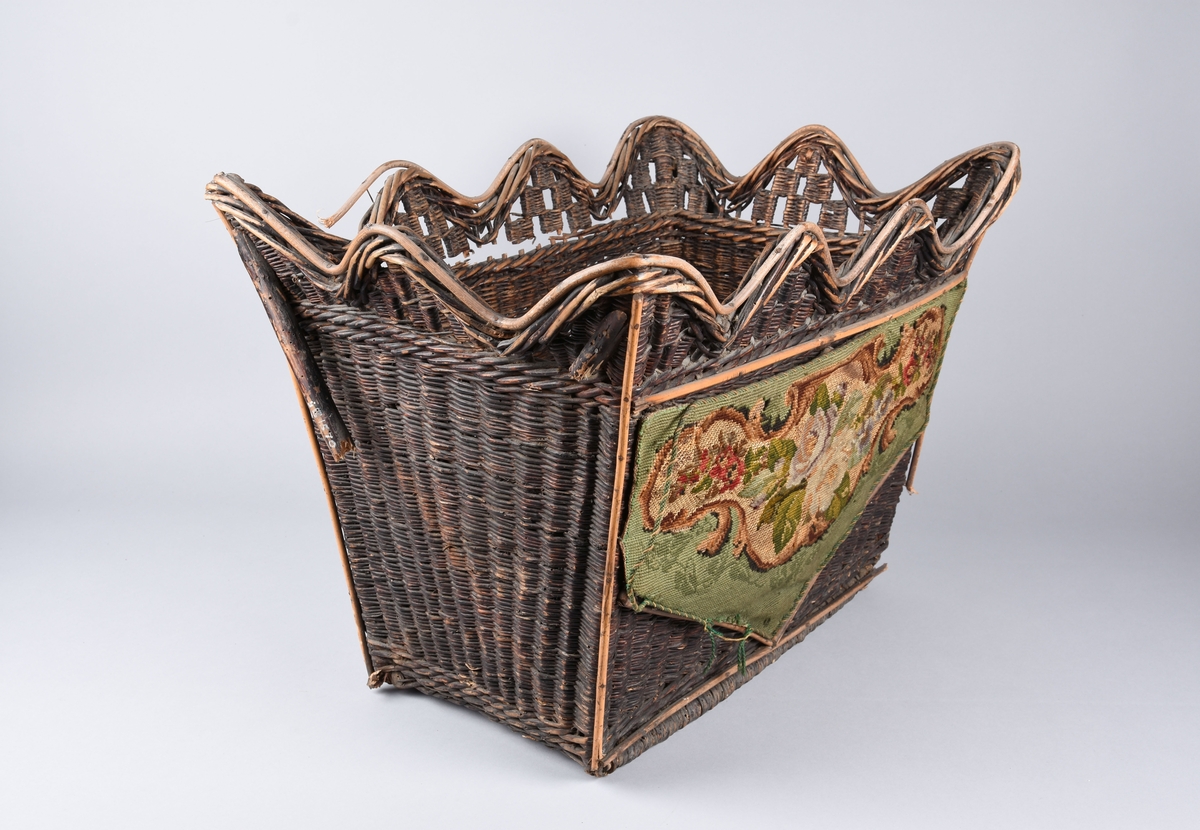 Flettet kurv av vidje med avstivere på tverrsidene. 
Brunmalt. Den ene langsiden er pyntet med broderi i ullgarn. Blomstermønster mot grønn bakgrunn.