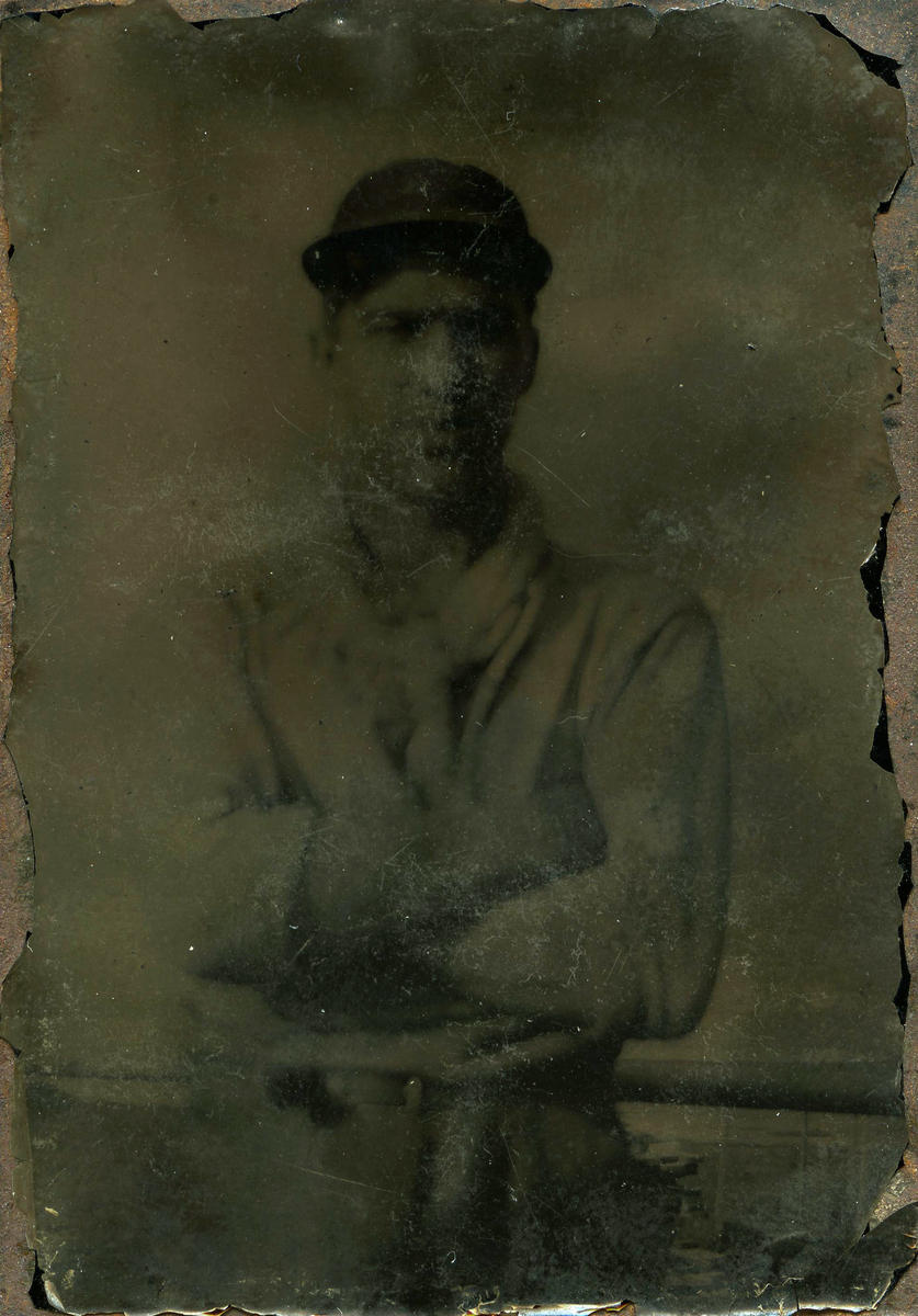 Fotografi på metallplate som viser en ung man som står ved et rekkverk med armene i kryss og en lue på hodet. Han har på seg en lys, langarmet skjorte, en skjerf og en bukse med belte. Bak rekkverket kan man se konturene av bebyggelse og kystlandskap.