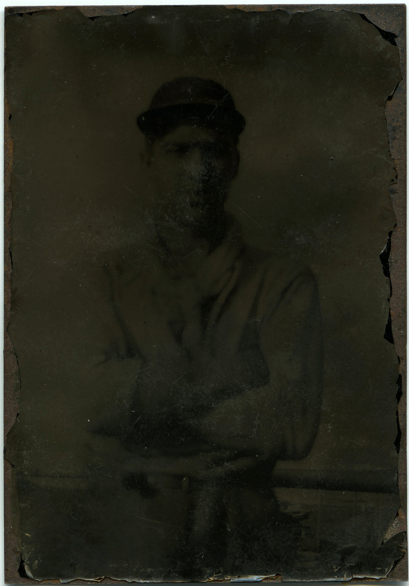 Fotografi på metallplate som viser en ung man som står ved et rekkverk med armene i kryss og en lue på hodet. Han har på seg en lys, langarmet skjorte, en skjerf og en bukse med belte. Bak rekkverket kan man se konturene av bebyggelse og kystlandskap.