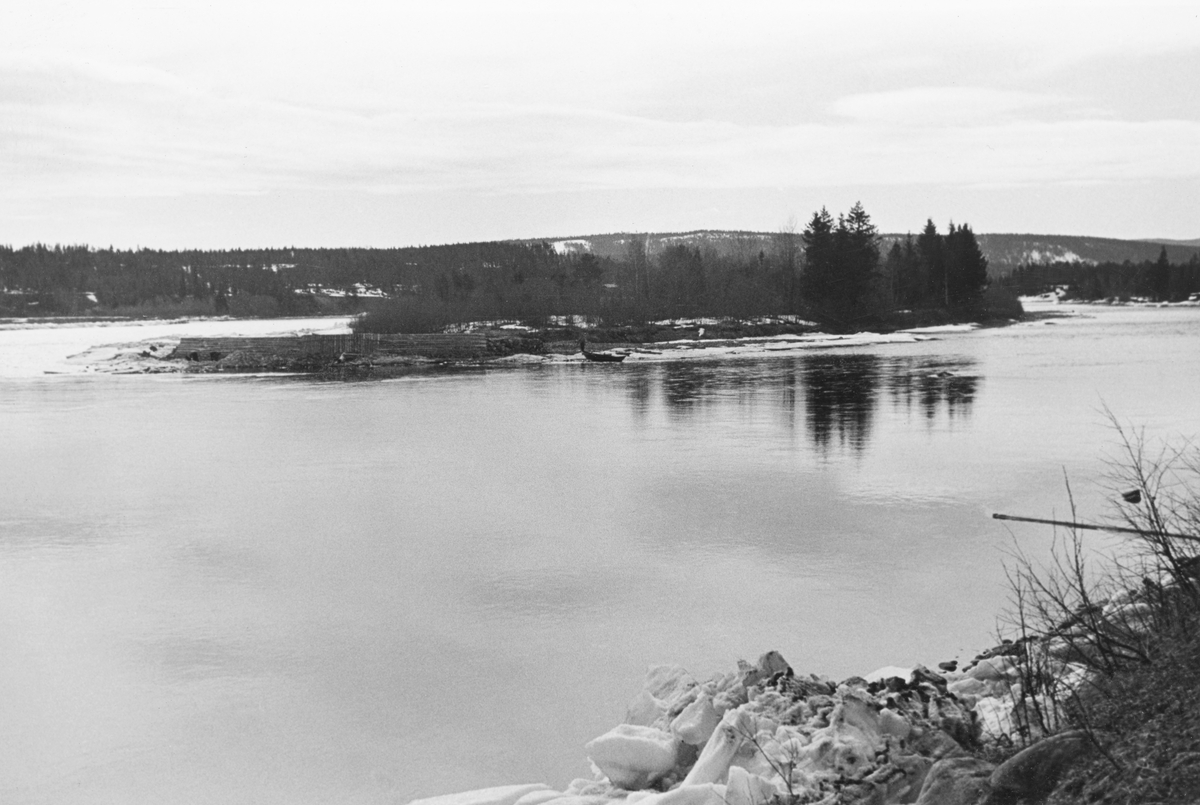 Dette fotografiet er registrert inn i Norsk skogmuseums fotodatabase med teksten «Glomma i Stor-Elvdal, trolig i området ved Stai». Det er slett ikke usannsynlig at dette motivet er fra Glomma i Stor-Elvdal, men årsaken til at dette motivet fantes i Glomma fellesfløtingsforenings arkiv var nok at det arbeidet som pågikk på motstrøms side av den lille flate øya i elveløpet da bildet ble tatt. Her ble det bygd en skråstilt skjerm av tre, så såkalt skådam, som, når våren kom og fløtinga startet, skulle lede tømmeret til side for øya. Vi skimter et par karer som arbeidet med fundamenteringa ved fronten av skådammen. Båten de tok seg til og fra arbeidsstedet med lå i øyas strandsone, til høyre på bildet. Også der skimtes en mann. Bildet skal være tatt i begynnelsen av mai 1939, mens vannføringa i Glomma ennå var lav nok til at det var mulig å utføre forbygningsarbeider.