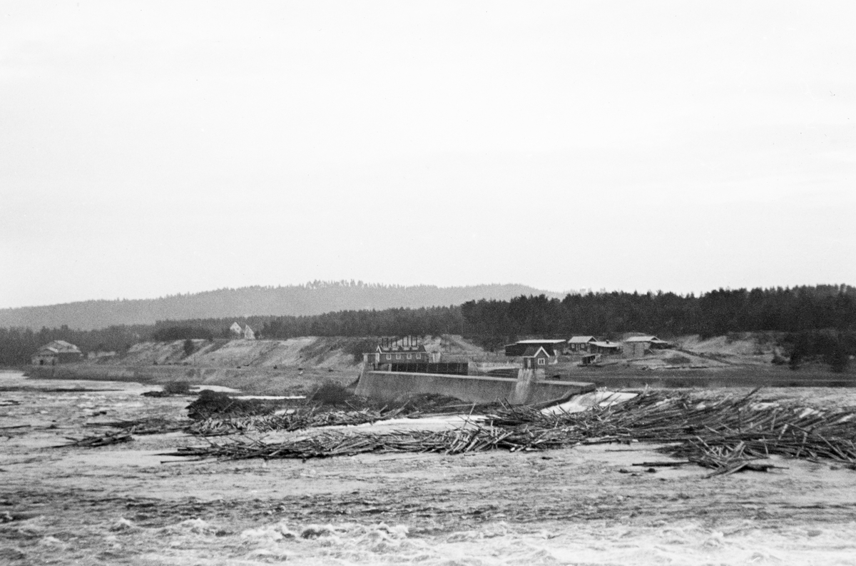 Tømmerhaug ved kraftverksdammen i Skjefstadfossen i Glomma, nærmere bestemt i Heradsbygda i Elverum, Hedmark. Fotografiet ble tatt i 1938, fra østre elvebredd langs den buete overløpsdammen. Detter var en 312 meter lang betongmur med avrundet krone som skulle stuve opp elvevannet og på den måten magasinere over 600 millioner kubikkmeter vann. Fløtingstømmeret på motstrøms side av dammen ble forsøkt ledet mot den østre elvebredden, for der var elveløpet djupest, noe som reduserte faren for at stokkene skulle sette seg fast og stuves sammen i hauger. Målet var at tømmeret kunne flyte uhindret videre nedover med strømmen. Da dette fotografiet ble tatt hadde det bygd seg opp en haug av tømmerstokker som sprikte i mange retninger under den vestre delen av overløpsdammen. Denne haugen stengte for det tømmeret som lå på den ovenforliggende dammen. Her var det krevende oppgaver som ventet på fløterne. Helt til venstre i bildet skimter vi Skjefstadfoss kraftsasjon.