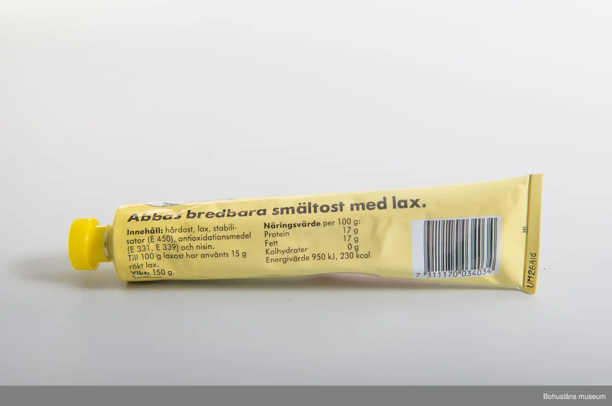 Tryckt dekor i gult,rosa,svart,vitt Gul gjuten plasthatt. Text: "Abbas Lax-Ost. Nyhet  samt produktbild  På baksidan innehållsförteckning och prismärkningskod. 
Tuben är identisk med UM026808
Om givaren se UM026667