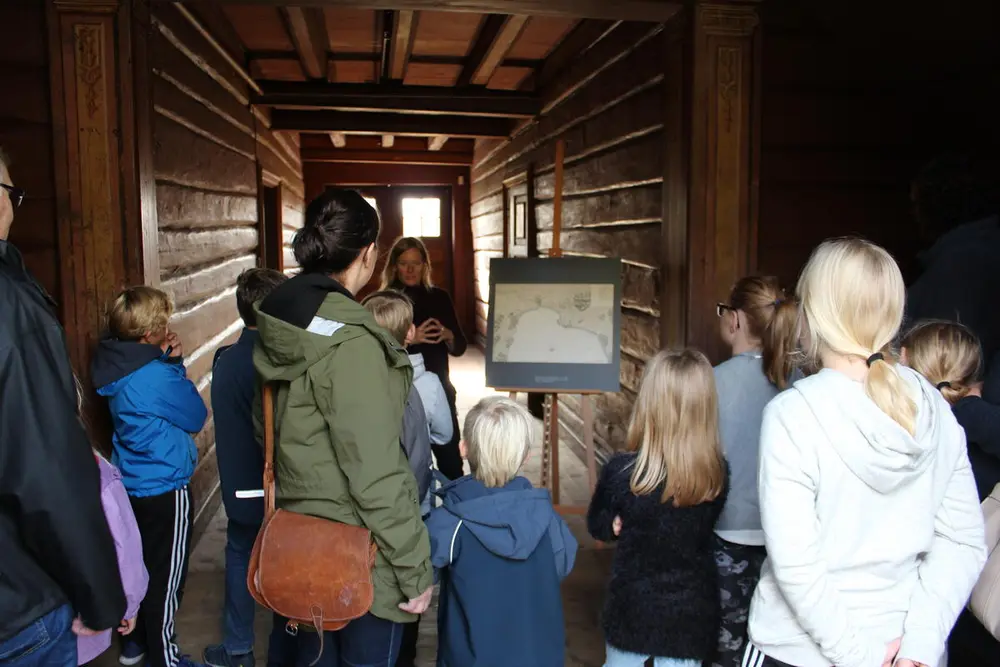 Flere mennesker står med ryggen til og hører på en som forteller. De er i et bygg med mørke tømmervegger