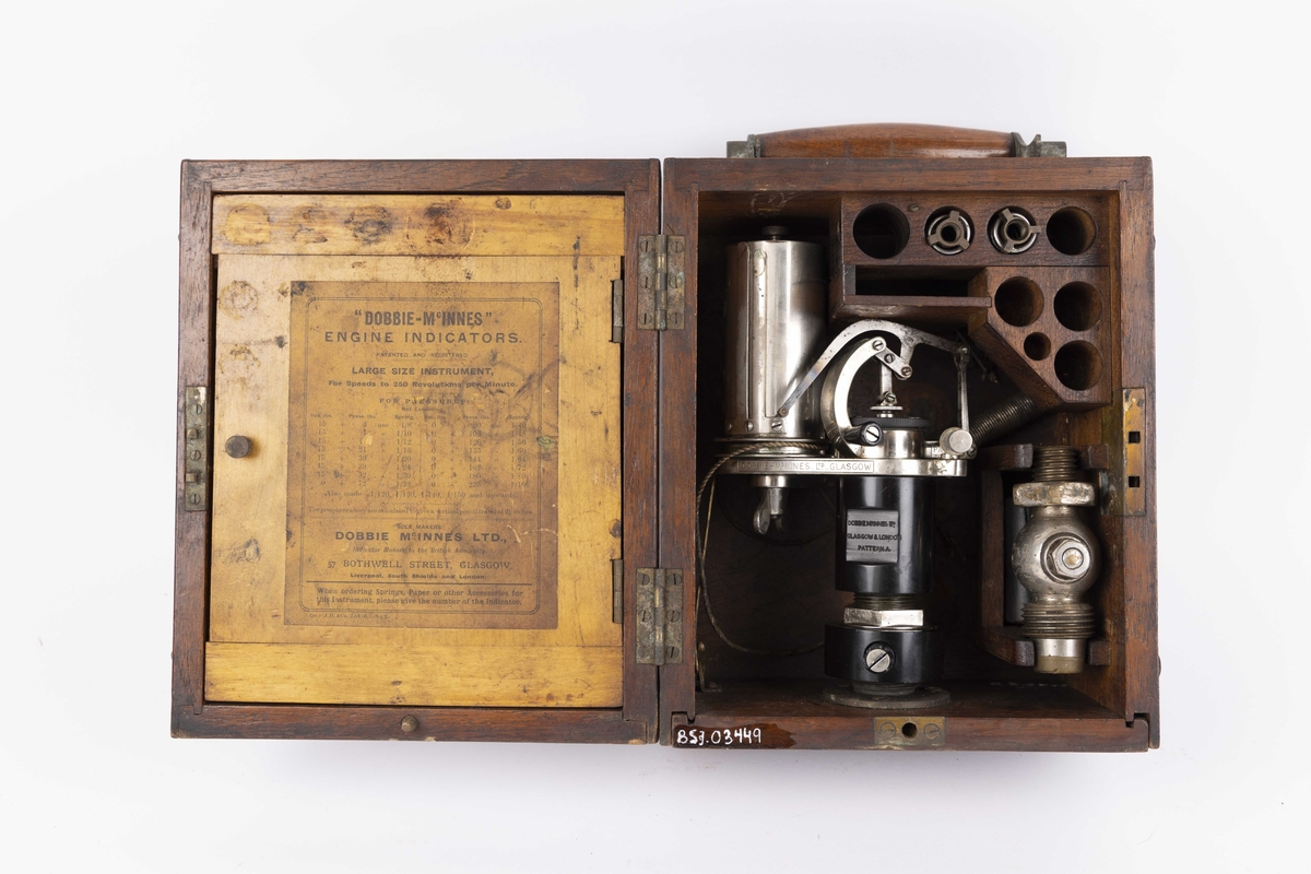 Dampmaskinindikator med flere deler i brun trekasse. Kassen har lokk med hengsler, innvendig hengslet plate til romavdeler og oppbevaringsfester for løse deler. Indikatoren er festet til ene kortsiden og vippes ut ved bruk. Instrumentet har en sylinder. 
