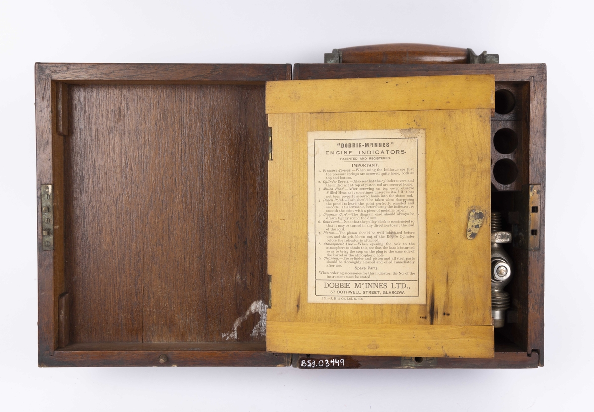 Dampmaskinindikator med flere deler i brun trekasse. Kassen har lokk med hengsler, innvendig hengslet plate til romavdeler og oppbevaringsfester for løse deler. Indikatoren er festet til ene kortsiden og vippes ut ved bruk. Instrumentet har en sylinder. 

