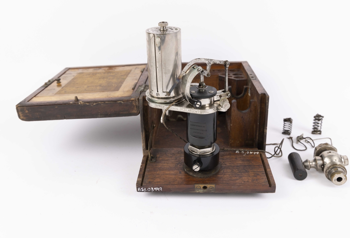 Dampmaskinindikator med flere deler i brun trekasse. Kassen har lokk med hengsler, innvendig hengslet plate til romavdeler og oppbevaringsfester for løse deler. Indikatoren er festet til ene kortsiden og vippes ut ved bruk. Instrumentet har en sylinder. 
