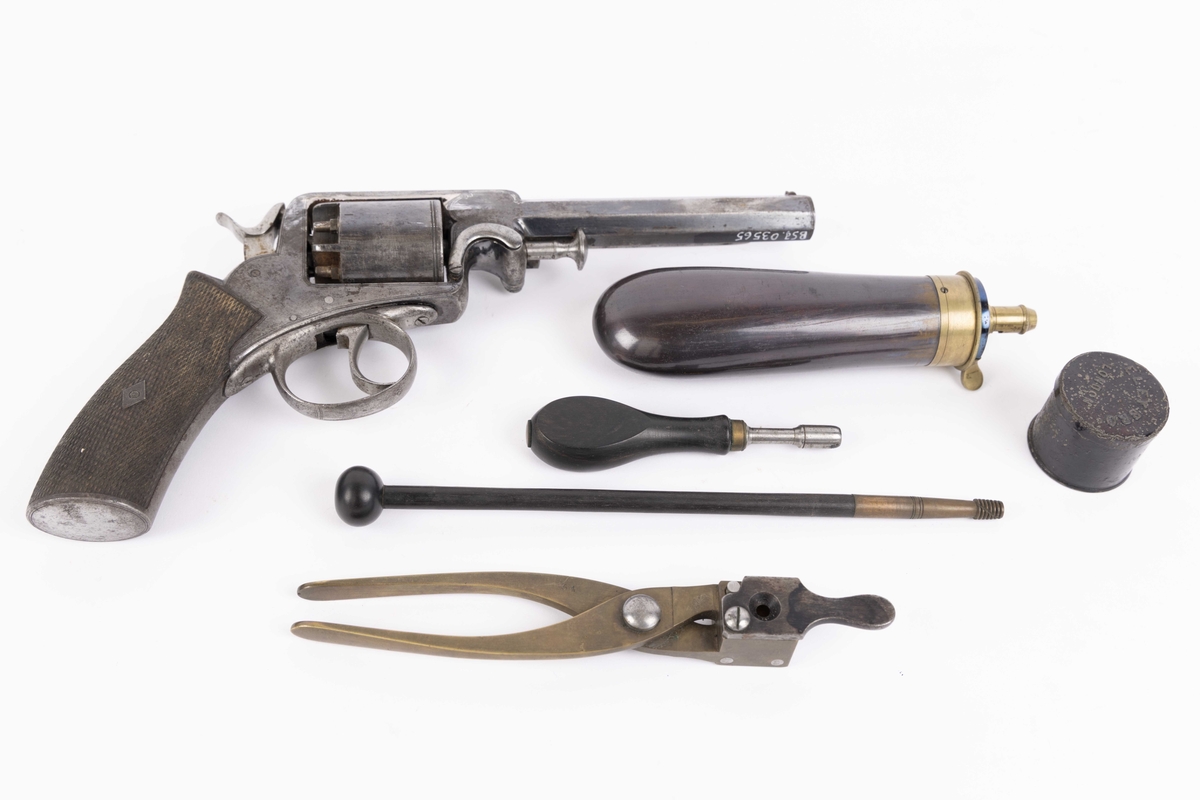 Revolver i treskrin.  Seks skudds Lee perkusjonsrevolver med krutthorn, kulestøpetang til forming av blykuler, demonteringsnøkkel for tennhettenippler, 6 blykuler, pussestav og metallboks inneholdende voks for smøring. I tillegg boks for oppbevaring av krutt (tømt for innhold).  Treetui av mahogny foret med blå filt innvendig. Mangler tennhetter.