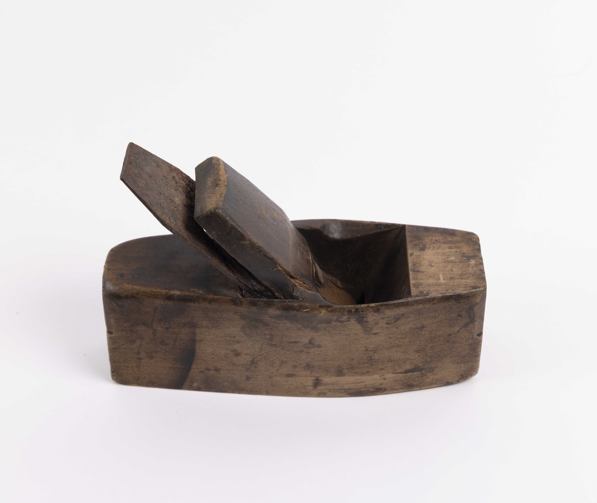 Rektangulær pusshøvel i tre med flat bunn og avrundede langsider. Den har et bredt høveljern som er kilt fast i høvelen med en trekile.
