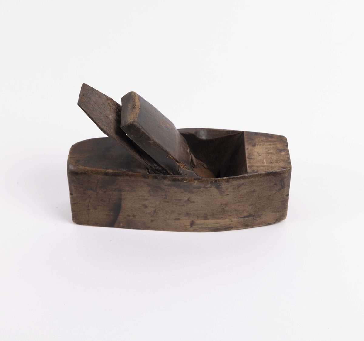 Rektangulær pusshøvel i tre med flat bunn og avrundede langsider. Den har et bredt høveljern som er kilt fast i høvelen med en trekile.
