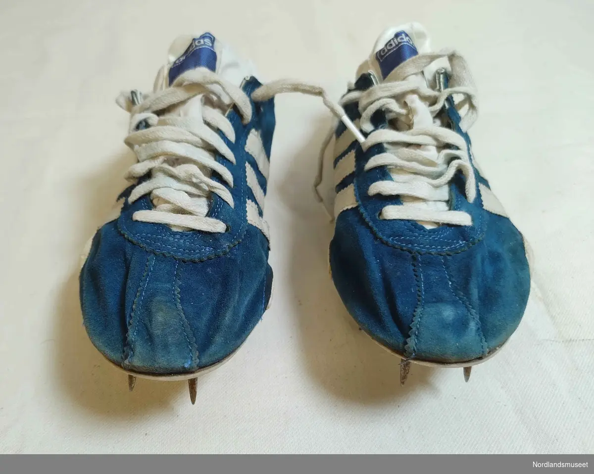 Et par idrettssko med pigger under sålen, og snøring. Antatt brukt til friidrett. Blå med hvite striper på sidene. Semsket skinn. I original skoeske som mangler lokk.