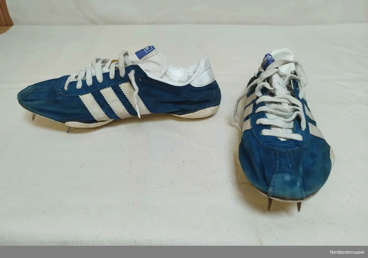 Et par idrettssko med pigger under sålen, og snøring. Antatt brukt til friidrett. Blå med hvite striper på sidene. Semsket skinn. I original skoeske som mangler lokk.