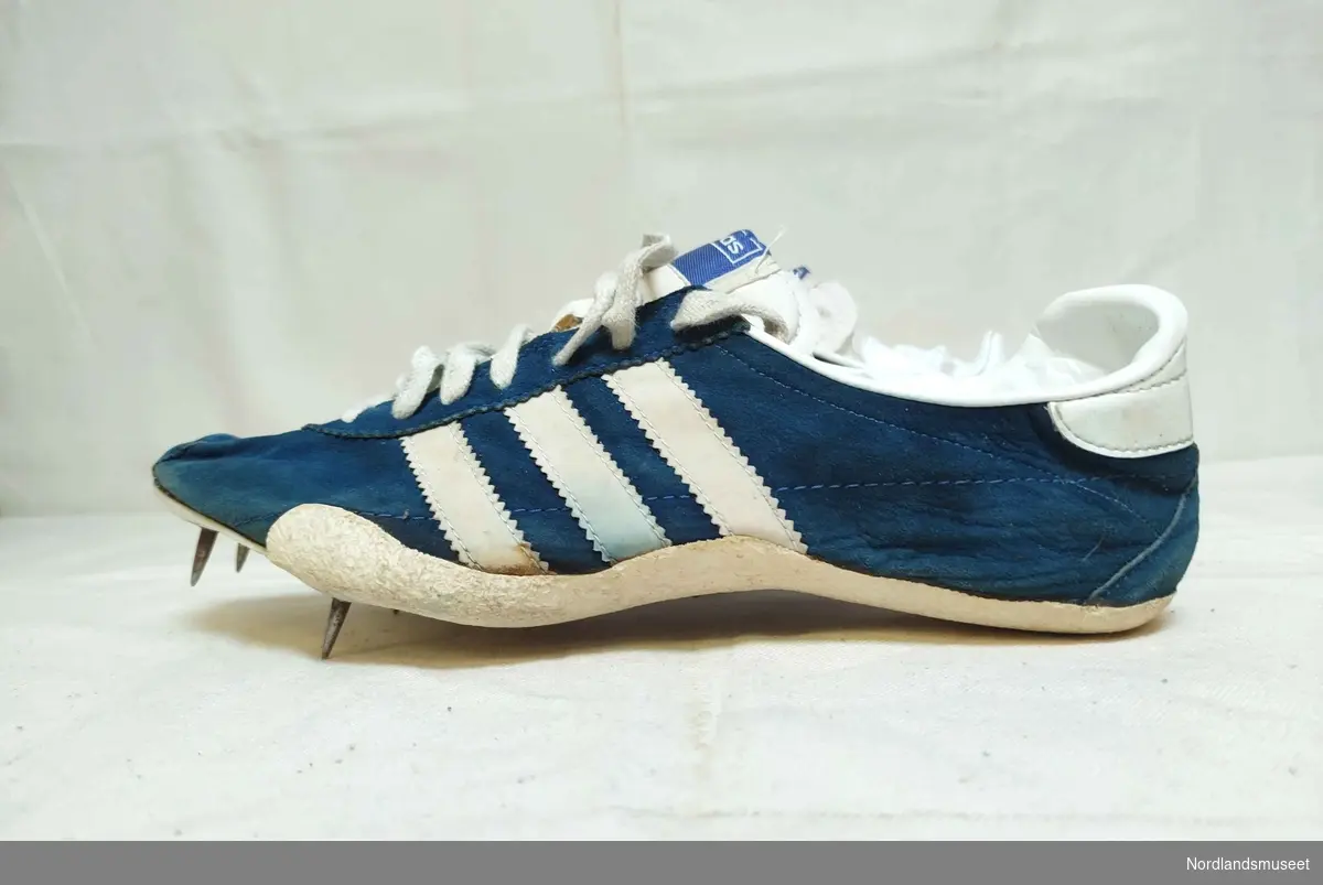 Et par idrettssko med pigger under sålen, og snøring. Antatt brukt til friidrett. Blå med hvite striper på sidene. Semsket skinn. I original skoeske som mangler lokk.