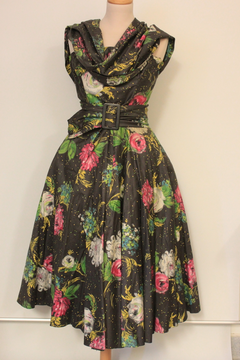 Klänning i tre delar samt skärp. Ärmlöst liv, lös schalkrage samt separat kjol. Tyget är grafitgrått med blommönster i rosa, vitt , gult och grönt. Flera stärkta underkjolar hör till. 