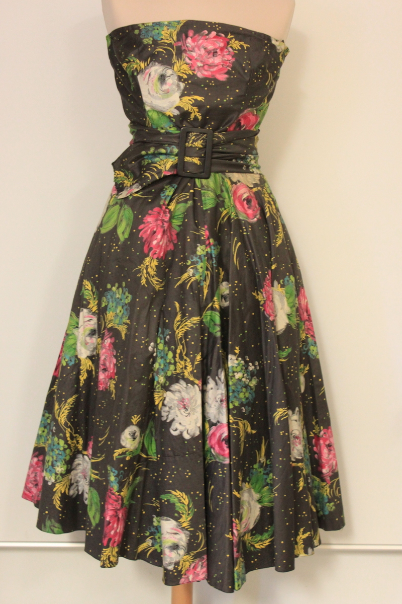 Klänning i tre delar samt skärp. Ärmlöst liv, lös schalkrage samt separat kjol. Tyget är grafitgrått med blommönster i rosa, vitt , gult och grönt. Flera stärkta underkjolar hör till. 