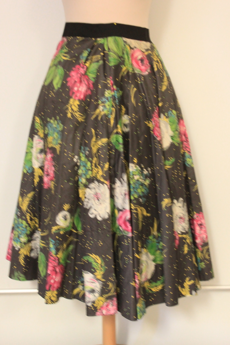 Klänning i tre delar samt skärp. Ärmlöst liv, lös schalkrage samt separat kjol. Tyget är grafitgrått med blommönster i rosa, vitt , gult och grönt. Flera stärkta underkjolar hör till. 