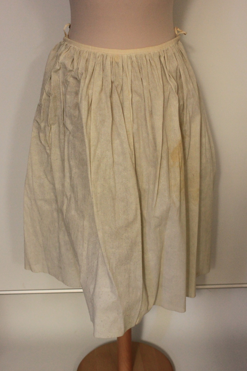 Klänning i tre delar samt skärp. Ärmlöst liv, lös schalkrage samt separat kjol. Tyget är grafitgrått med blommönster i rosa, vitt , gult och grönt. Flera stärkta underkjolar hör till. 