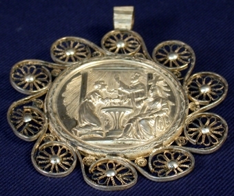 Cirkelrund medaljong, 58 mm i diameter, runtom dekorerad med böjda silverband med tenntråd formad till blomma, inuti. En silverögla sitter på kanten, igenom vilken en kedja eller liknande kan träs så att medlajongen kan hängas. På medaljongens framsida ett bildmotiv i relief: I tempelliknande byggnad med kolonner knäböjer en kvinna och håller fram sitt nyfödda barn för dop i en dopfunt. En man i skägg och särk döper barnet, medan en sittande kvinna  i särk och slöja på huvudet betraktar. Hon håller i ett kors. Bredvid henne finns symbolerna ankare och hjärta. Strålglans kommer från himlen. På medaljongens baksida tre stycken stämplar.