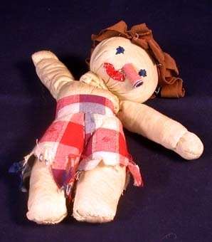 Trasdocka av tyg. Hemmasydd. Dockkropp av beige tyg på vilken sytts fast brunt tyghår i form av breda tygremsor, blå ögon, cylinderformad, utstående näsa, röd mun och byxor från midjan ner till knäna i röd/vit/blå-rutigt tyg. Byxorna fransiga nedtill. Sömmar markerar händernas början. Extra tygremsa runt hals som stöd åt huvudet. På dockans bröst står skrivet "ASTRID".