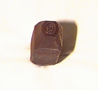 Rektangulärt metallstycke med avsmalnande ändar. I ena änden finns en platt slagyta, och i andra änden finns en inramad text i relief: "UB".