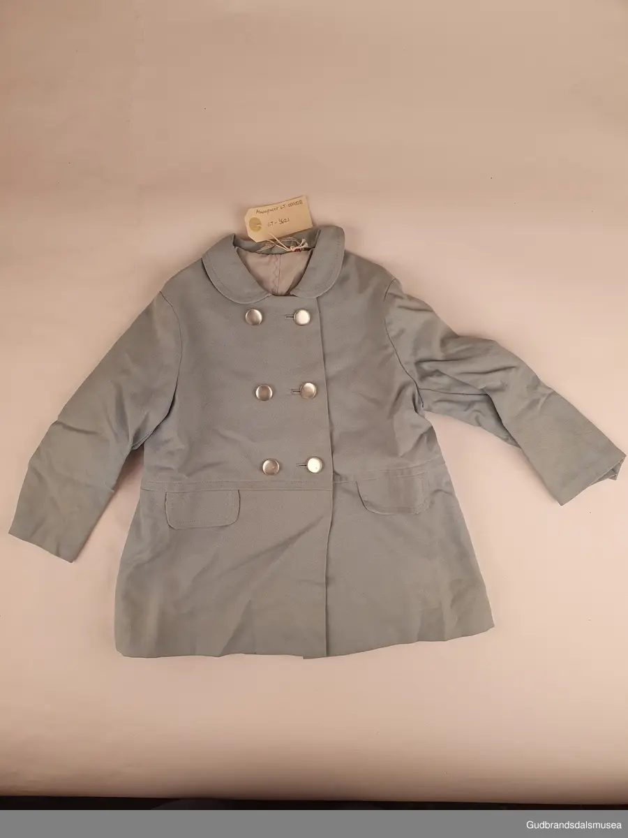 Hjemmesydd barnekåpe med tilhørende hatt. Dobbeltspent lukning, og overlappende splitt nede bak. Både kåpen og hatten er tynt lyseblått ullstoff , og er foret med forsilke. 2 lommeklaffer, påsydd kun til pynt. 6 stk hvite/blanke knapper i lukning foran. Hatten er i modell som en sydvest. med knapp i toppen.