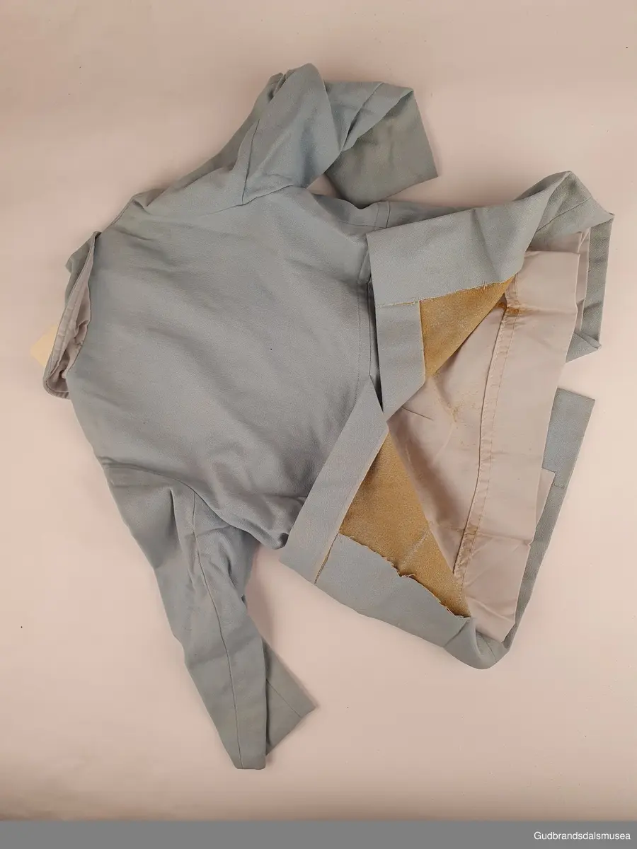 Hjemmesydd barnekåpe med tilhørende hatt. Dobbeltspent lukning, og overlappende splitt nede bak. Både kåpen og hatten er tynt lyseblått ullstoff , og er foret med forsilke. 2 lommeklaffer, påsydd kun til pynt. 6 stk hvite/blanke knapper i lukning foran. Hatten er i modell som en sydvest. med knapp i toppen.