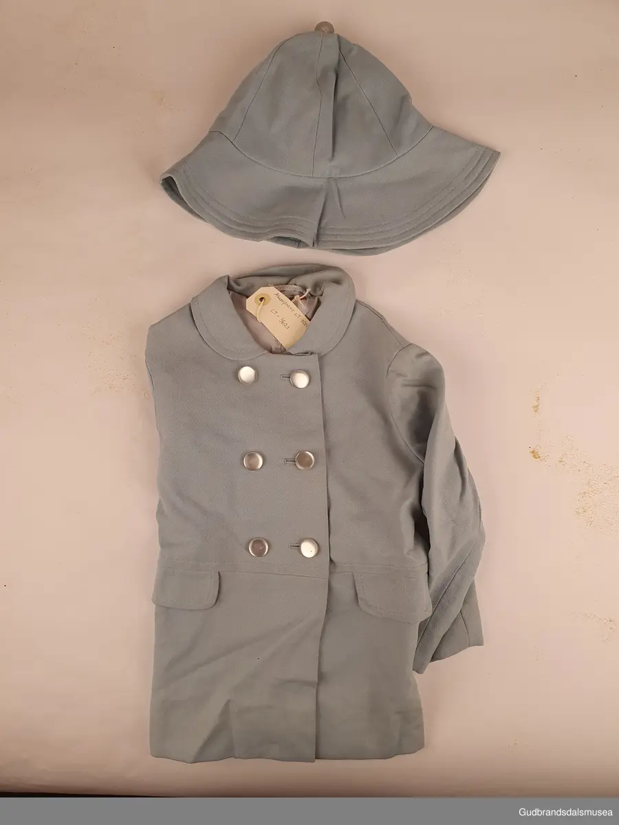 Hjemmesydd barnekåpe med tilhørende hatt. Dobbeltspent lukning, og overlappende splitt nede bak. Både kåpen og hatten er tynt lyseblått ullstoff , og er foret med forsilke. 2 lommeklaffer, påsydd kun til pynt. 6 stk hvite/blanke knapper i lukning foran. Hatten er i modell som en sydvest. med knapp i toppen.