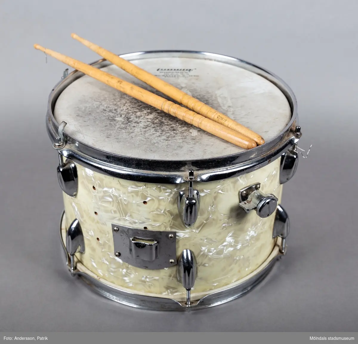 En rund trumma i metall och plastliknande material i cremefärgat pärlemoliknande mönster, samt två tillhörande avsmalnande långa trumpinnar i trä. Trumman ser ut att vara väl använd då ovansidan är missfärgadav trumslag. Trumman har ägts och spelats av Holger Åkerström. Holger var född 15/3 1904 i Örgryte.