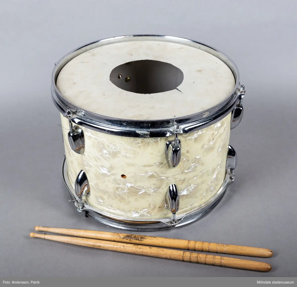 En rund trumma i metall och plastliknande material i cremefärgat pärlemoliknande mönster, samt två tillhörande avsmalnande långa trumpinnar i trä. Trumman ser ut att vara väl använd då ovansidan är missfärgadav trumslag. Trumman har ägts och spelats av Holger Åkerström. Holger var född 15/3 1904 i Örgryte.