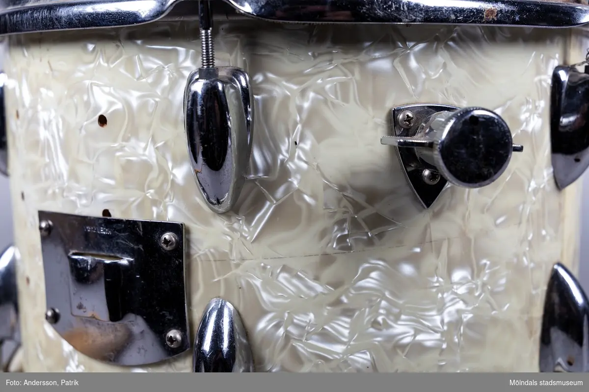 En rund trumma i metall och plastliknande material i cremefärgat pärlemoliknande mönster, samt två tillhörande avsmalnande långa trumpinnar i trä. Trumman ser ut att vara väl använd då ovansidan är missfärgadav trumslag. Trumman har ägts och spelats av Holger Åkerström. Holger var född 15/3 1904 i Örgryte.