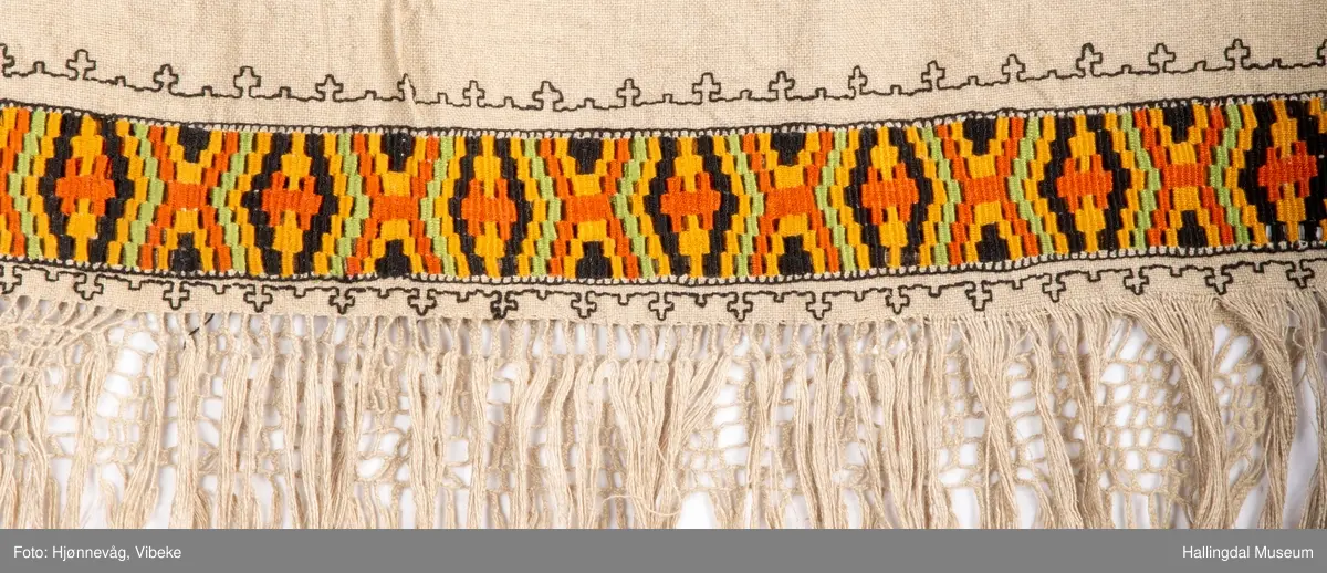 Vevd løper, toskaft i lin med frynser, beige. Broderi holbeinsøm. Nær frynsene er det broderi som ser ut som det er kelimvevd (uttrekksøm); svart, orange, sennep og grønt. Frynser med firflettet blonde i begge ender. Firflettingen er ikke gjort ferdig.