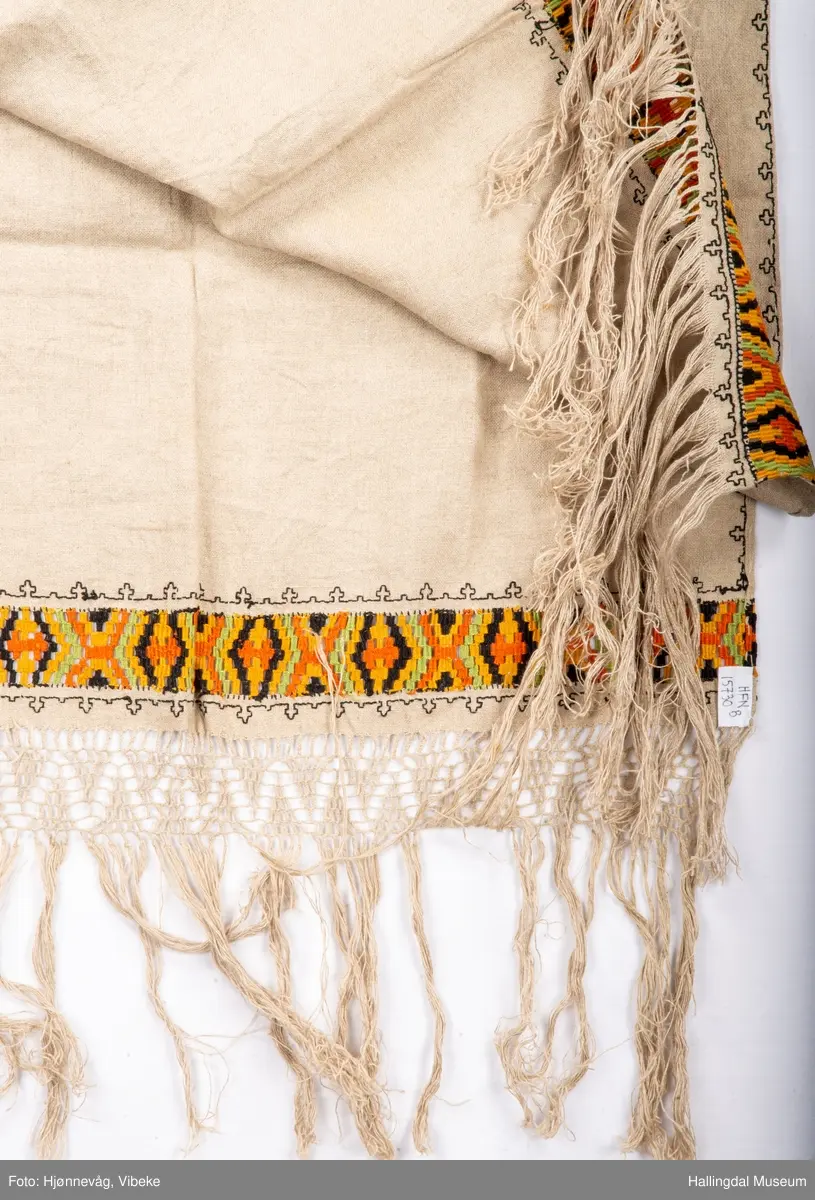 Vevd løper, toskaft i lin med frynser, beige. Broderi holbeinsøm. Nær frynsene er det broderi som ser ut som det er kelimvevd (uttrekksøm); svart, orange, sennep og grønt. Frynser med firflettet blonde i begge ender. Firflettingen er ikke gjort ferdig.